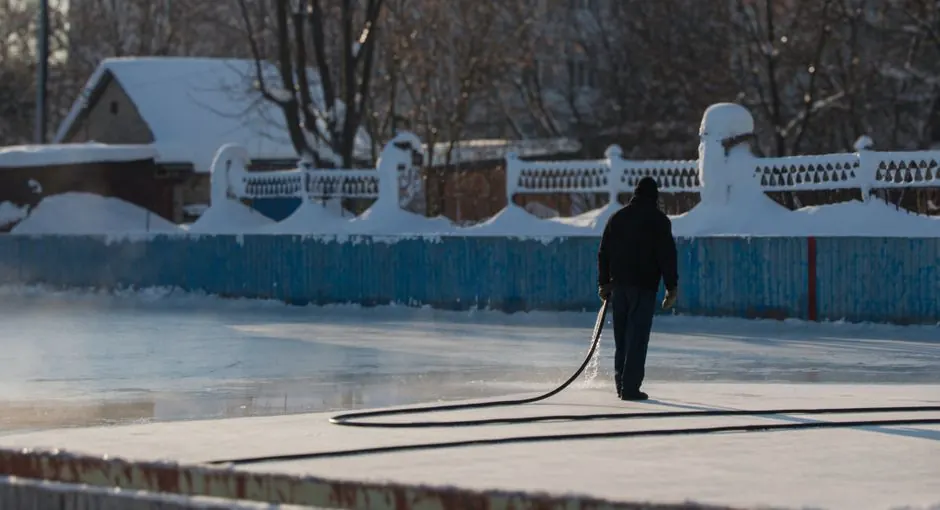 Person using a hose to maintain an outdoor ice rink during winter with snow in the backdrop.